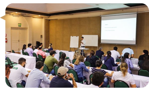 Pablo Hurtado dando una conferencia en un salón con varias mesas y asistentes tomando notas. Una pantalla proyecta diapositivas con información relevante y un pizarrón está ubicado junto al conferencista.